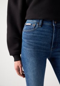 Tamno plave traperice izrađene od denima, s srednjim strukom, dizajnom s pet džepova i bijelom oznakom Calvin Klein blizu pojasa.