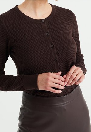 Femme portant un cardigan boutonné texturé marron foncé et une jupe en cuir marron foncé, les mains jointes au niveau de la taille.