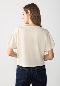 T-shirt crème coupé avec des manches courtes ; tissu lisse et encolure ronde ; associé à un jean bleu foncé avec poches arrière.