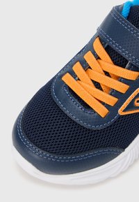 Tamsiai mėlynas tinklinis sportbačiai su oranžinėmis raišteliais, paminkštinta liežuviu ir balta padu. Yra kontrasto siūlės ir brendintas akcentas šone.