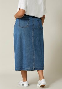 Jupe en denim de longueur mi-mollet en bleu moyen. Présente deux poches arrière, une coupe droite et des coutures minimales. Associée à des baskets blanches.