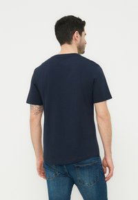 Άντρας με σκούρα μαλλιά, που φοράει ένα απλό σκούρο μπλε μπλουζάκι και μπλε τζιν, δείχνοντας την πλάτη του μπροστά σε απλό λευκό φόντο.