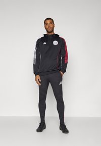 Sudadera deportiva negra con media cremallera, rayas rojas y blancas en las mangas y un logo. Combinada con leggings negras a juego y zapatillas deportivas.