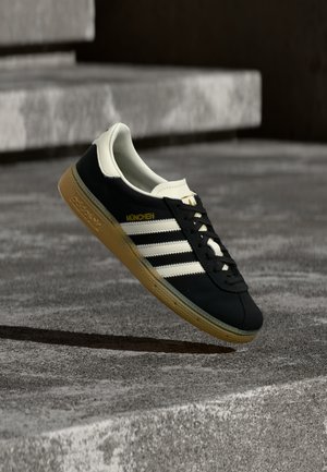 Black Adidas München sneaker with white stripes and gum sole hovering above textured concrete steps.