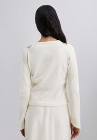 Haut crème à manches longues avec un tissu texturé, présentant un large décolleté et des découpes aux épaules, conçu pour une coupe décontractée.