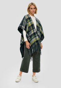 Geruite poncho in groen, marineblauw en grijs; met franjes aan de zoom; gedragen over een witte longsleeve en olijfgroene broek.