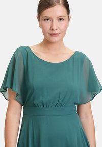 Türkises Kleid mit transparenten, schwingenden Ärmeln und gerafftem Bund. Glattes Material mit lockerer Passform. Rundhalsausschnitt und leichte Textur.
