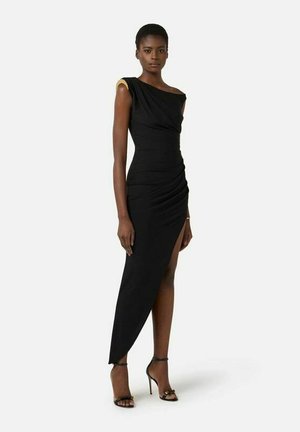 Donna che indossa un abito nero monospalla con uno spacco alto e dettaglio dorato sulla spalla, abbinato a sandali neri con tacco alto.