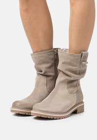 Bottes en suède beige à la cheville avec un design froncé, une fermeture éclair sur le côté et une semelle texturée beige clair. Aucun motif ni accent supplémentaire n'est visible.