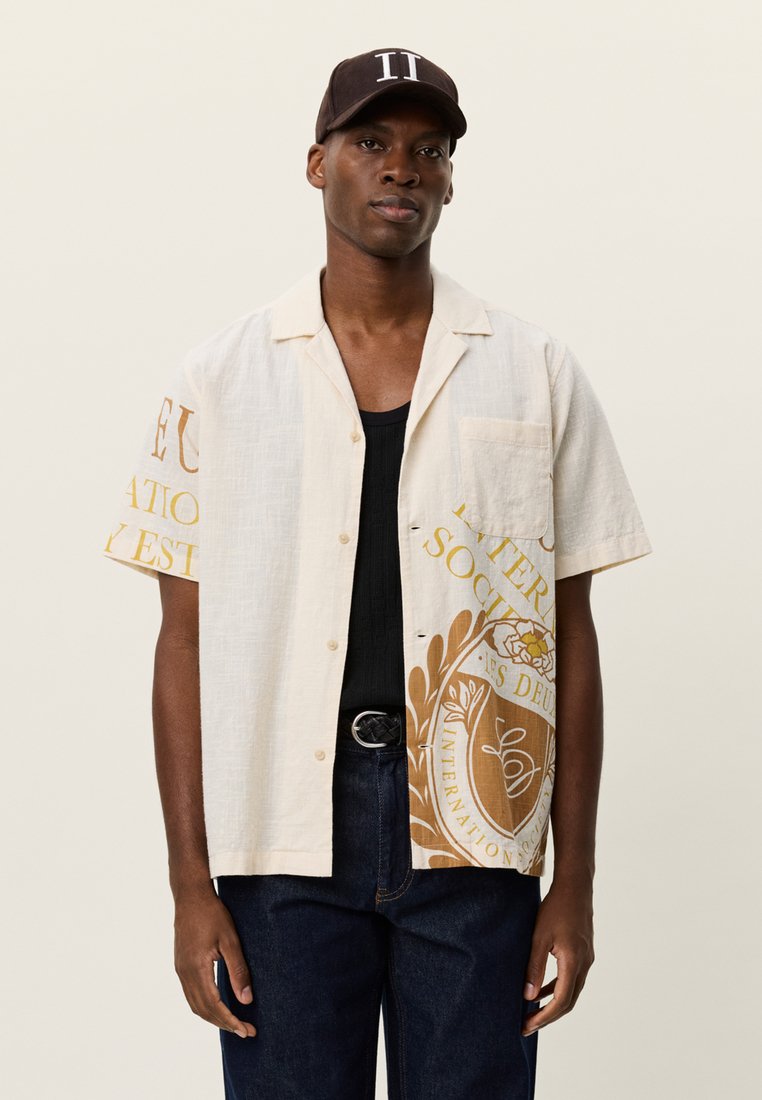 Πουκάμισο από λινό με κοντά μανίκια σε κρεμ χρώμα με κίτρινο και καφέ γραφικό τύπωμα. Διαθέτει τσέπη και άνετη γραμμή, συνδυασμένο με σκούρο τζιν.