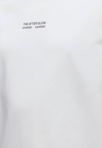 Λευκό ύφασμα με μικρό μαύρο κείμενο "THE AFTER GLOW SAMSØE SAMSØE" κοντά στο πάνω κέντρο