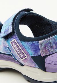 Sandal azul y púrpura con un tejido resistente al agua, correa ajustable y suela texturizada. Presenta un logo de Geox y acentos de costura.