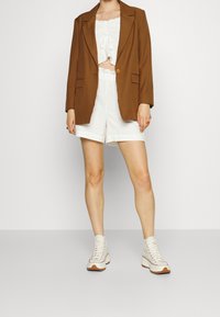 Brun kavaj, vit kort topp med dragsko, vita shorts och krämfärgade höga sneakers. Slät tyg, avslappnad passform, minimal detaljering.