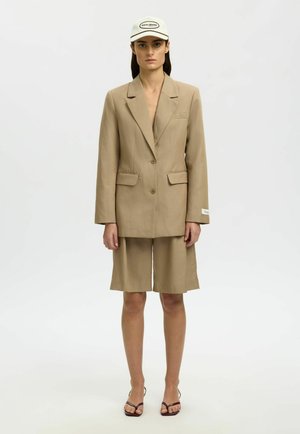 Donna con blazer beige oversize e pantaloncini abbinati fino al ginocchio, cappellino bianco e sandali marroni con tacco e punta aperta, in piedi davanti a uno sfondo neutro.