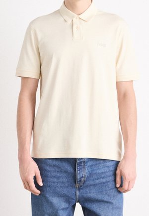 Homme portant un polo à manches courtes crème avec deux boutons et un jean bleu, debout devant un fond blanc uni.