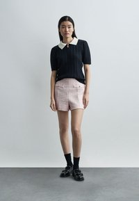 Femme portant un haut court en tricot noir à manches courtes avec col blanc, un short rose texturé, des chaussettes noires et des mocassins noirs, debout devant un mur gris uni.
