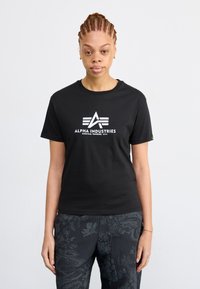 T-shirt en coton noir à manches courtes, arborant le logo et le design graphique "ALPHA INDUSTRIES" en blanc. Associé à un pantalon texturé à motif sombre.