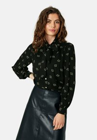 Blusa nera con fiocco, caratterizzata da un motivo geometrico in oro. Le maniche lunghe e la chiusura con bottoni completano la gonna in pelle elegante.