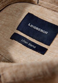 Etiqueta de mezcla de lino en una tela de color beige claro. La etiqueta superior dice "LINDBERGH," y la etiqueta inferior indica "Mezcla de Lino" en blanco sobre un fondo azul marino.