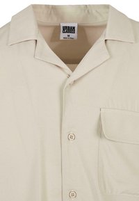 Chemise beige à boutons avec un col et une poche à rabat sur le côté gauche, étiquetée Urban Classics, taille moyenne, fabriquée en Chine.