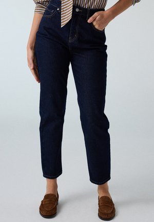 Donna che indossa jeans a vita alta blu scuro con mano in tasca e mocassini in camoscio marrone, in piedi davanti a uno sfondo semplice.