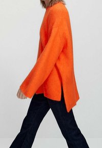 Pull en tricot orange à manches longues, avec fentes latérales et coupe décontractée, assorti à un jean foncé. Texture douce avec une apparence douillette.