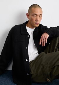 Cardigan nero lavorato a maglia con un ampio colletto a costine e bottoni argentati, indossato sopra una camicia bianca. Pantaloni verdi visibili, in posa seduta contro un muro bianco.