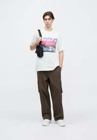 Fehér grafikás póló lila szöveggel és felhőképpel, laza barna cargo nadrággal párosítva. Kis fekete válltáskát tart. Fehér sportcipő.