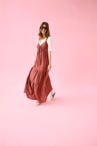 Femme avec des lunettes de soleil portant une robe longue couleur rouille par-dessus un top blanc, marchant sur un fond rose avec des baskets blanches.