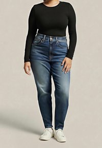 Personne portant un jean bleu taille haute, un haut noir à manches longues et des baskets blanches, se tenant devant un fond beige uni.