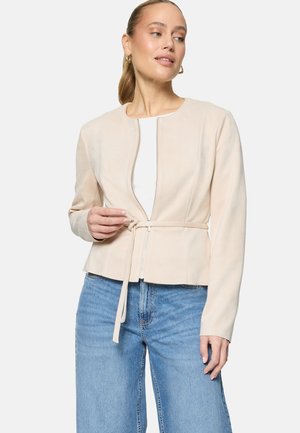 Frau, die eine beige Reißverschlussjacke mit Gürtel über einem weißen Oberteil anpasst, dazu hellblaue Jeans und goldene Creolen trägt.