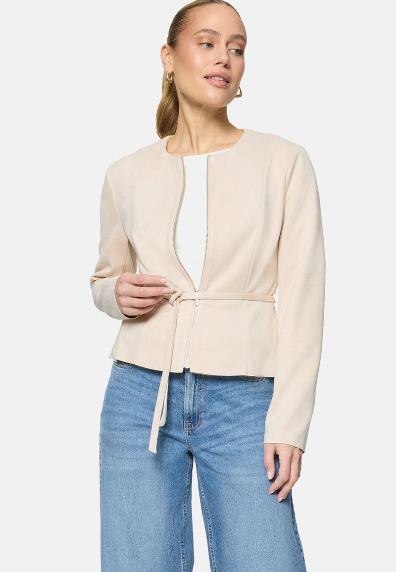Femme ajustant une veste beige zippée avec ceinture sur un haut blanc, portant un jean bleu clair et des boucles d'oreilles anneaux dorés.