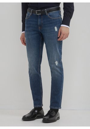 Mężczyzna ubrany w niebieskie postrzępione jeansy, czarny pasek, czarną koszulę i czarne skórzane mokasyny stoi na jednolitym tle.