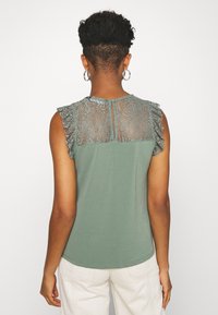 Zielony bezrękawnik z koronkowym tyłem, falbanami na ramionach oraz zapięciem na guzik przy szyi, wykonany z miękkiego, lekkiego materiału.