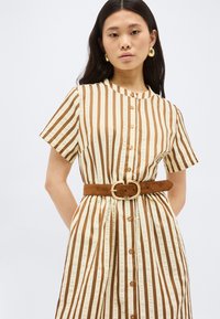 Γυναίκα που φοράει μπεζ και καφέ κάθετα ριγέ φόρεμα με κουμπιά, κοντά μανίκια και φαρδιά καφέ ζώνη με στρογγυλή αγκράφα.