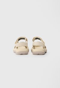Beige sandaalit, joissa on teksturoitu yläosa, säädettävät hihnat ja pehmustettu pohja, pyöreä kärki ja minimalistinen muotoilu.