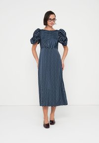 Robe marine à pois blancs, manches courtes bouffantes, corsage ajusté et jupe midi évasée. Portée avec des chaussures bordeaux.