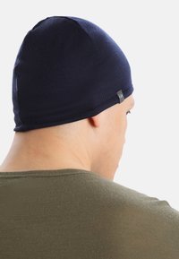 Navy modrá pletená čepice s těsným střihem, která má hladkou Texturu a malou logovou nášivku na straně, nošená osobou.