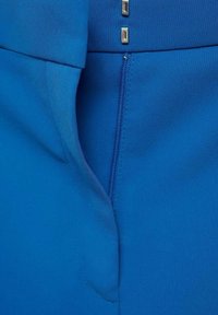 Gros plan d'un vêtement bleu montrant une fermeture éclair partiellement ouverte et deux crochets argentés au niveau de la ceinture.