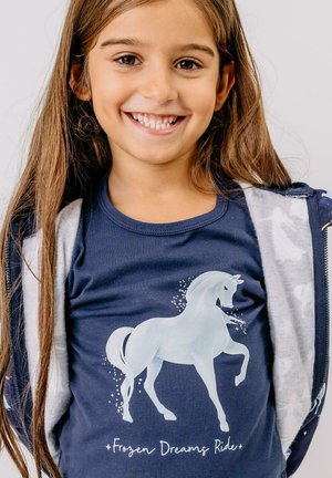 Marineblaues T-Shirt mit einem weißen Einhorn-Graphic und dem Text "Frozen Dreams Ride". Langes Haar und eine graue Zip-up-Jacke sind sichtbar.