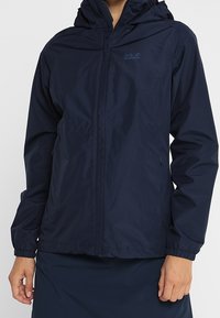 Marinblå vattentät jacka med huva, utrustad med frontdragkedja, elastiska ärmslut och diskret logotillverkning på bröstet. Slät textur.