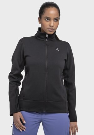 Frau trägt eine schwarze Jacke mit Reißverschluss und hohem Kragen sowie blaue Hosen, steht vor einem schlichten hellgrauen Hintergrund.