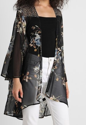 Woman wearing a sheer black floral kimono over a black top and white pants, showing flowing sleeves and open front design.