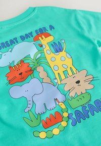 Türkisfarbenes kurzärmeliges Hemd mit cartoonartigen Tieren: einer gelben Giraffe, einer roten Katze, einem blauen Elefanten, einem grünen Krokodil und dem Text "GROSSARTIGER TAG FÜR EINE SAFARI."