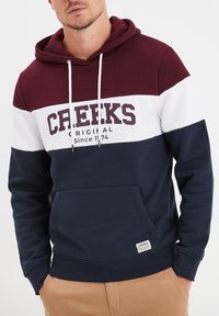 Felpa a righe bordeaux, bianca e blu navy in misto cotone. Caratterizzata da una tasca frontale e un cappuccio con lacci. Logo "CREEKS" al centro.