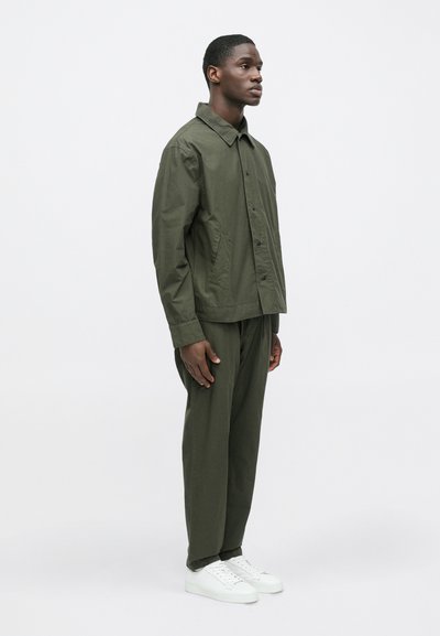 Olivovozelená bunda s gombíkovou légou a špicatým golierom, doplnená o matching nohavice a biele tenisky, zobrazená na neutrálnom pozadí.