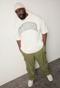 Weiße Kapuzen sweatshirt mit "STHUGE" Logo, kombiniert mit olivgrünen Cargo-Hosen und weißen Sneakern, stehend vor einem schlichten Hintergrund.