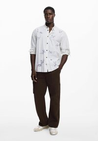 Gestreiftes Langarmshirt in Weiß mit blauen Designs, kombiniert mit lockeren braunen Cargo-Hosen und beigen Sportschuhen mit weißen Akzenten.