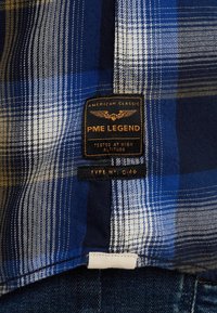 Tissu à carreaux bleu, blanc et beige avec une étiquette noire PME Legend portant la mention "American Classic, Tested at High Altitude, Type N° C-46."