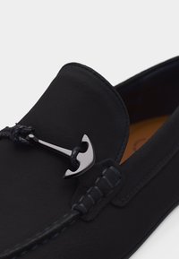 Mocassin en cuir noir avec détail tressé et ornement métallique en forme d'ancre argentée sur le dessus, sur un fond uni.